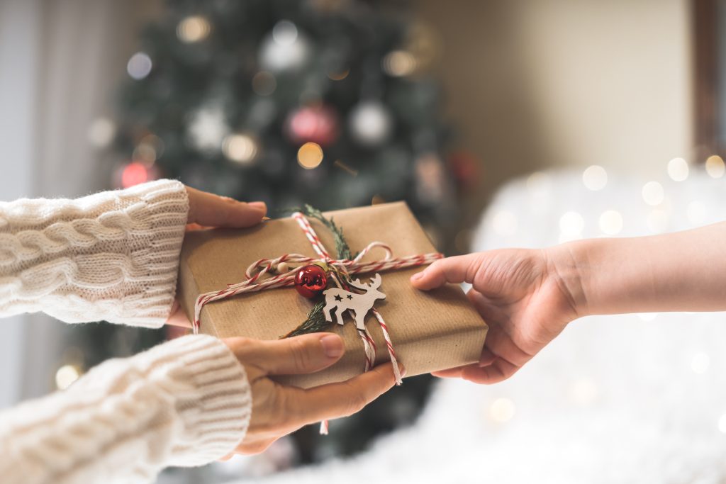 Woman In Sweater Giving A Wrapped Christmas Gift Box To Child. Glowing Snow Bokeh, Fir Tree. Winter Holidays